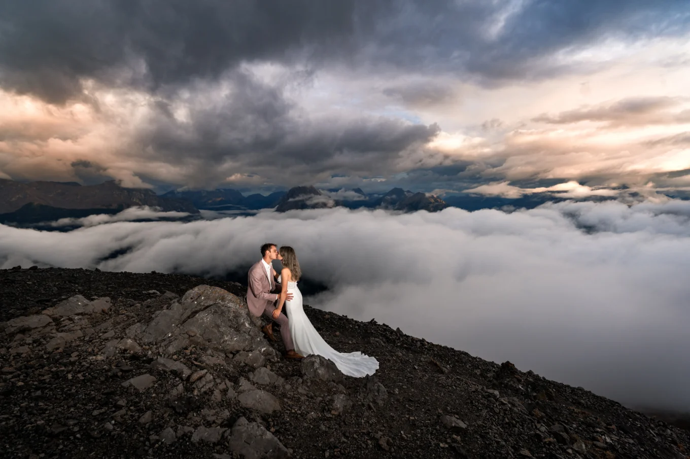 The bride kisses the groom who sits on the rock at the top on the mountain during coloreful sunset in Banff.