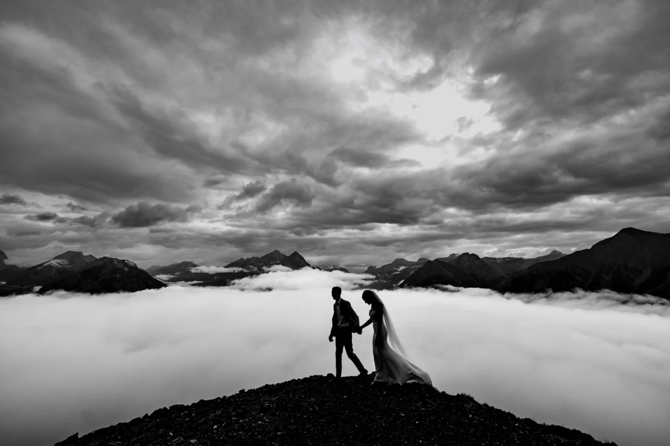 A couple in wedding attire walks triumphantly on a mountain peak, embracing each other with joy and love.