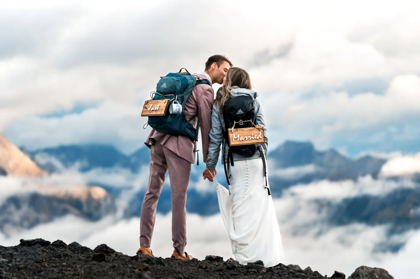 Bride and groom share a kiss at the summit of the Canadian Rockies wearing hiking backpacks.