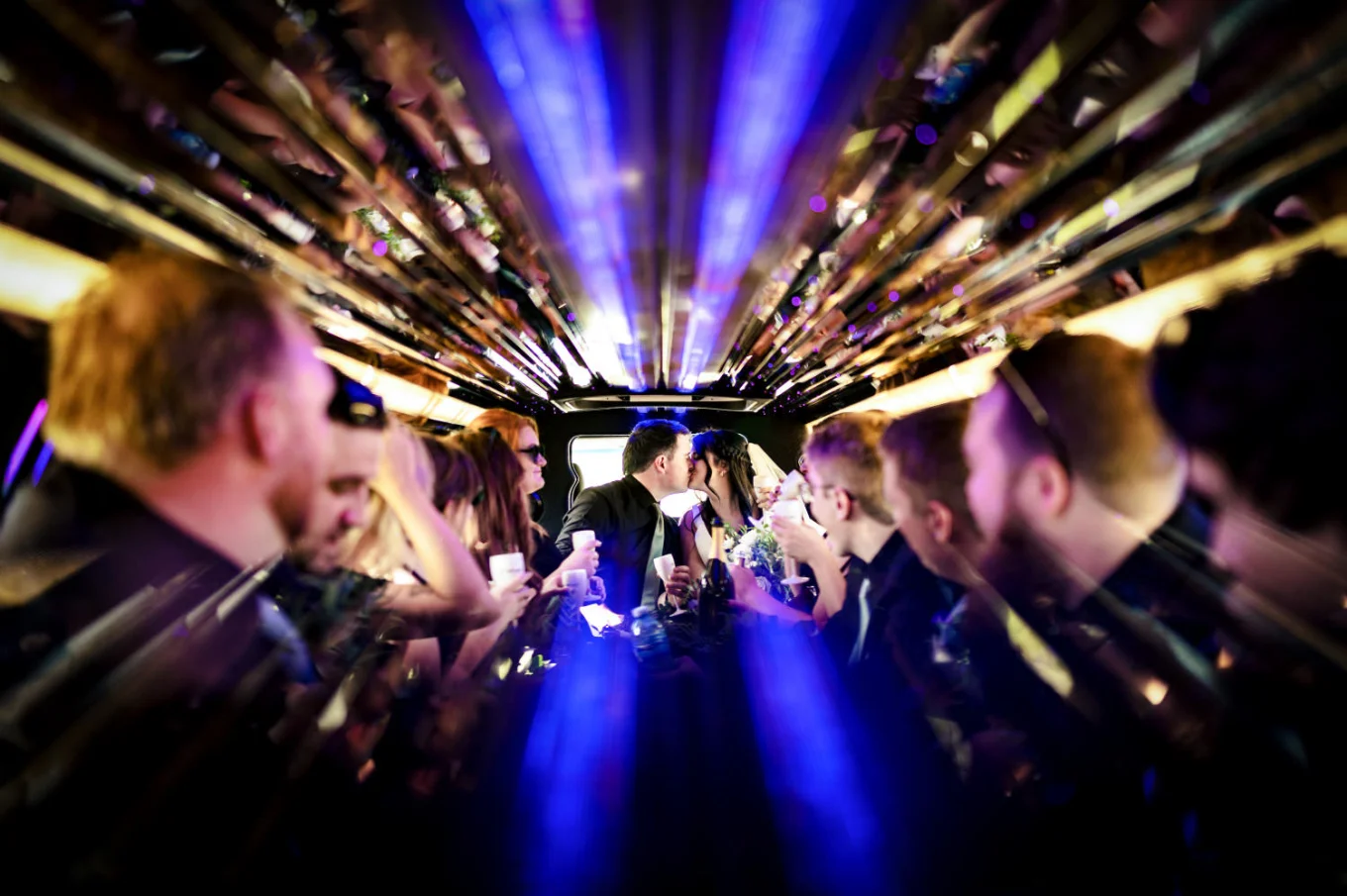 Newlyweds share a kiss in the limo while wedding party ookin at them