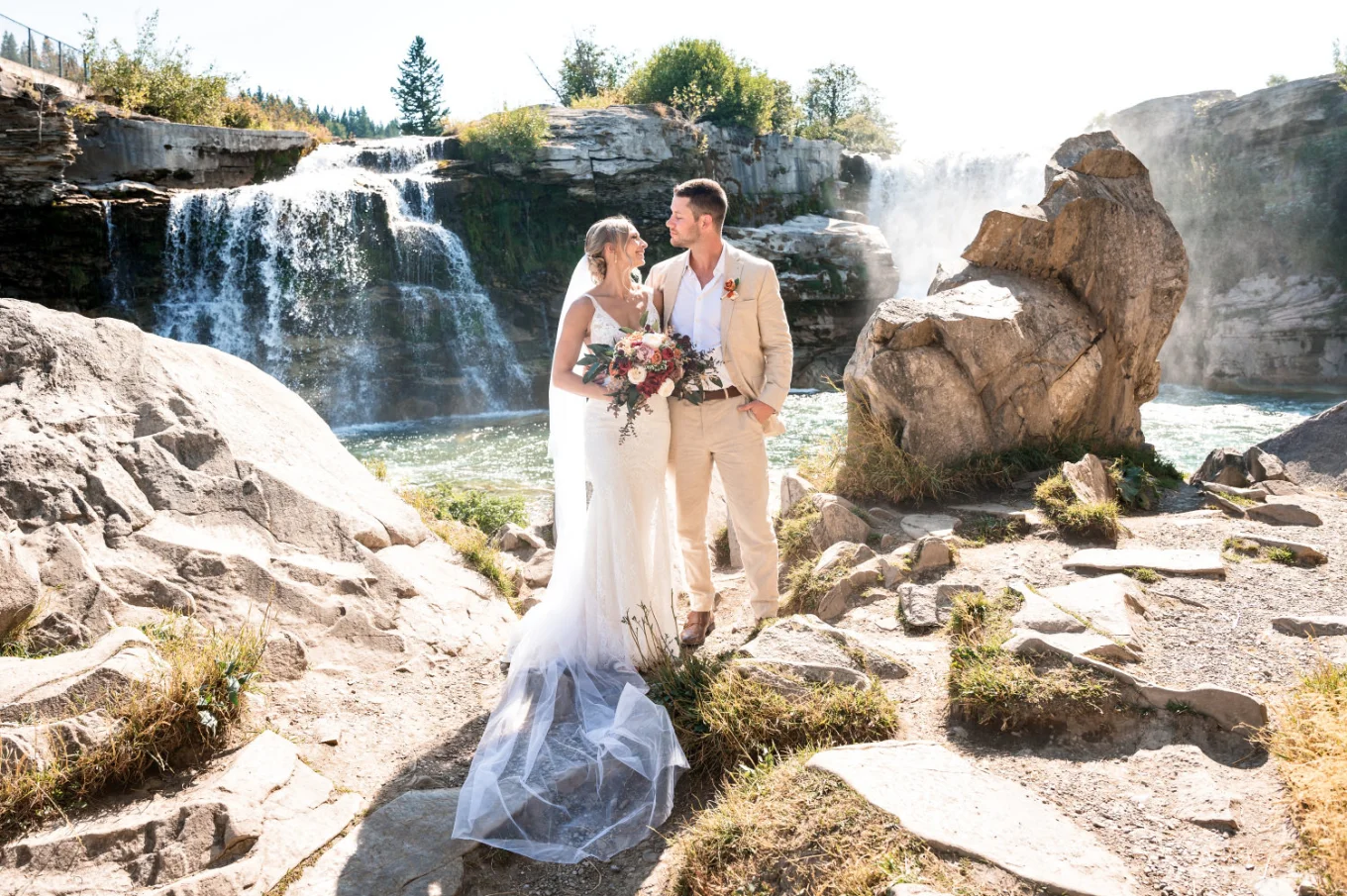 Bride an groom stand next other against stunning waterfall in Waterton.
