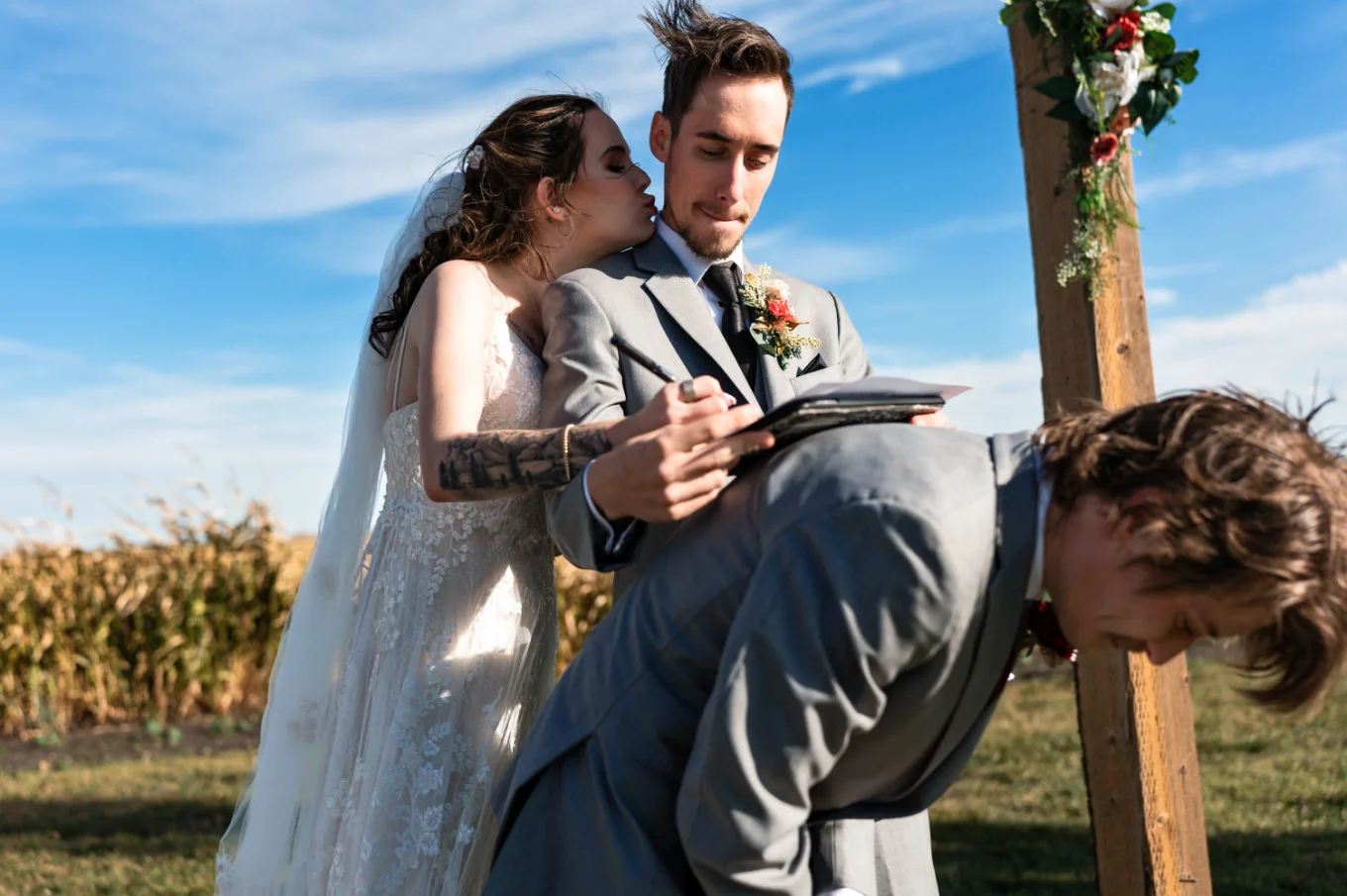 Groom sgh the wedding papers on the best-men back while the bride kisses him on the cheek.