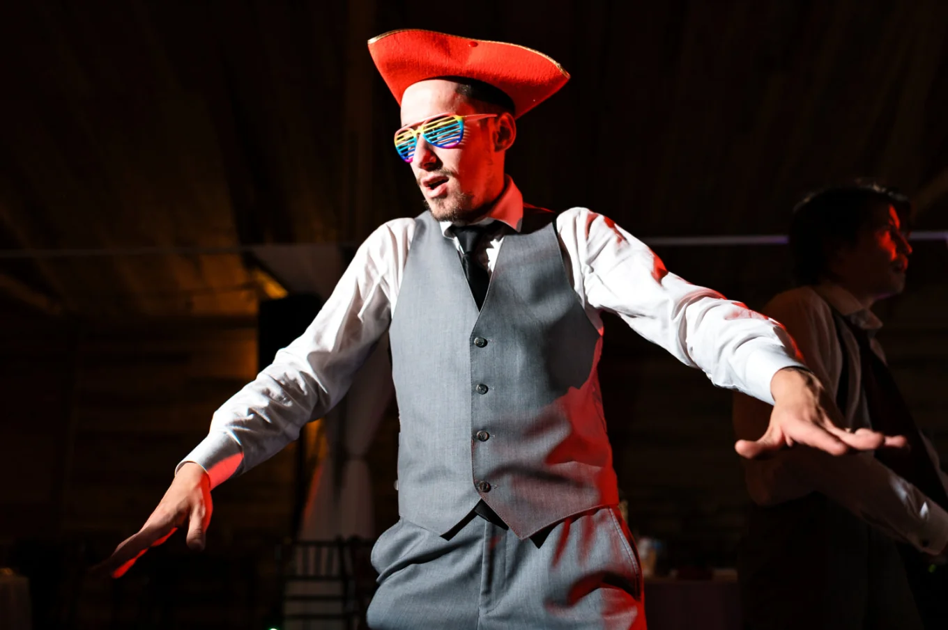 The groom wears a red, funny hat and glasses and dances