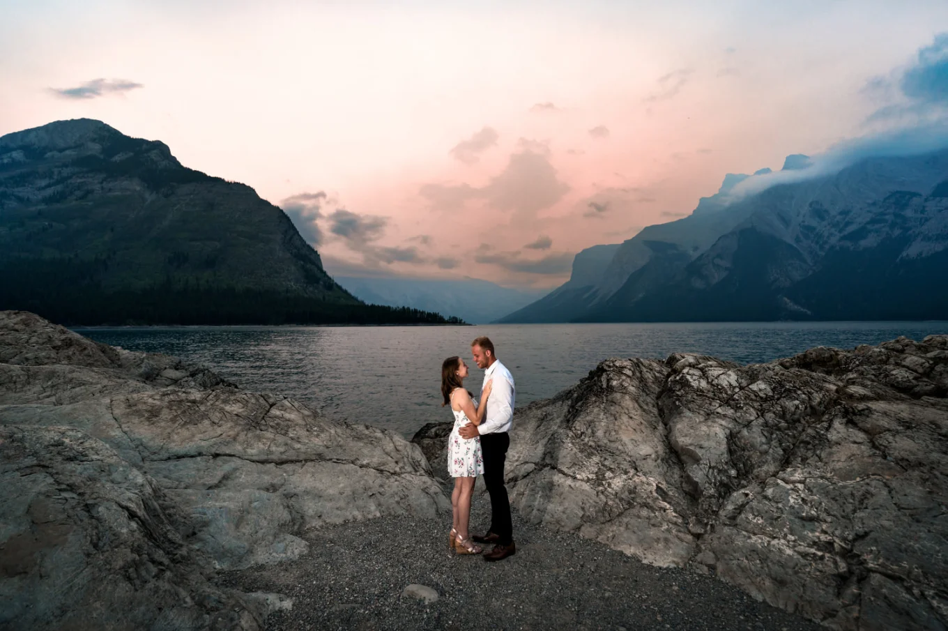 Engaged couple facing each other at Minnewanka Lake during colorefu sunrise