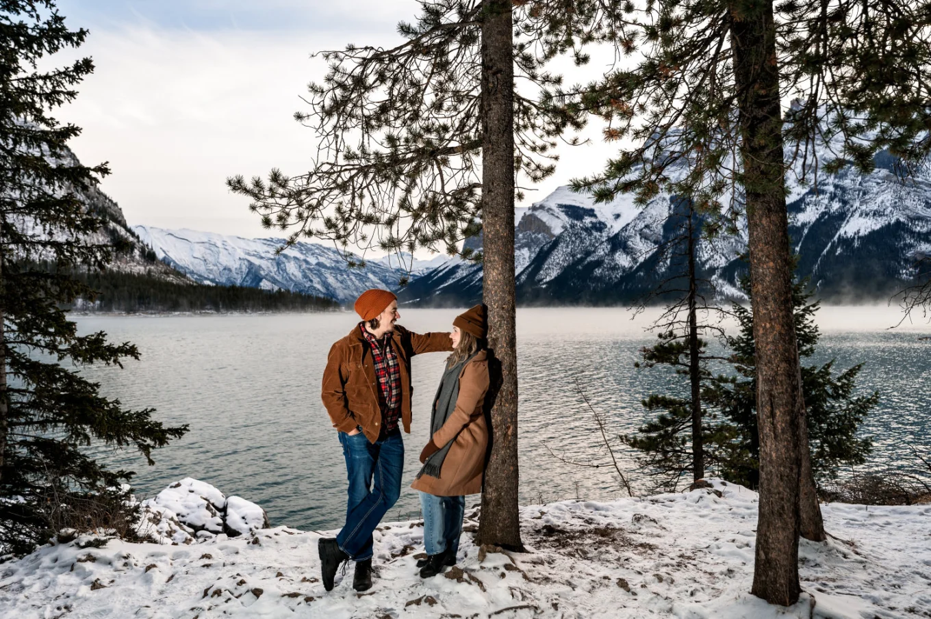 Lady leans over a tree at Minnewanka Lake while her fiancee look at her with love.