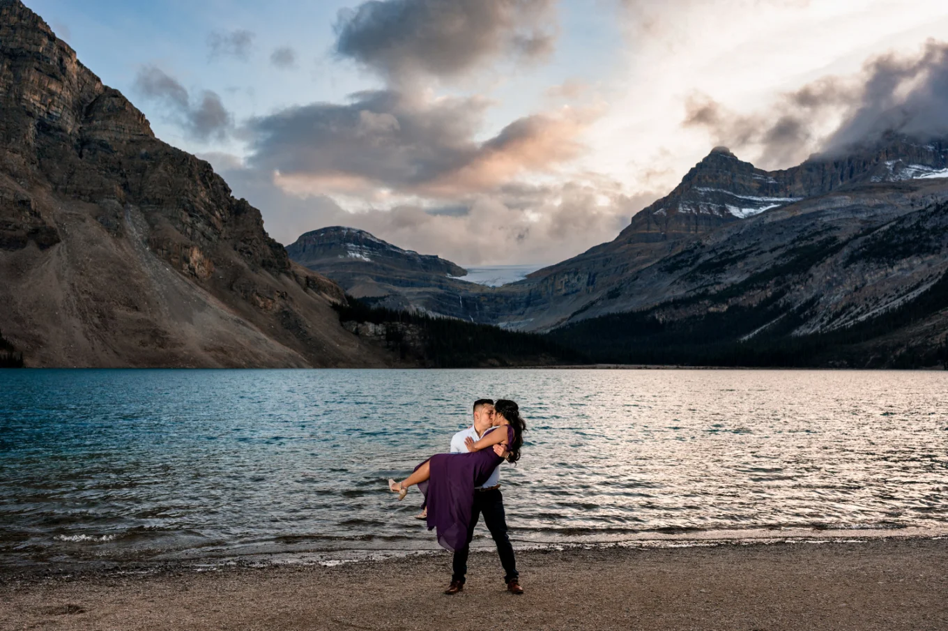 Man lifts his fiancee up at Bow Lake against huge mountains.
