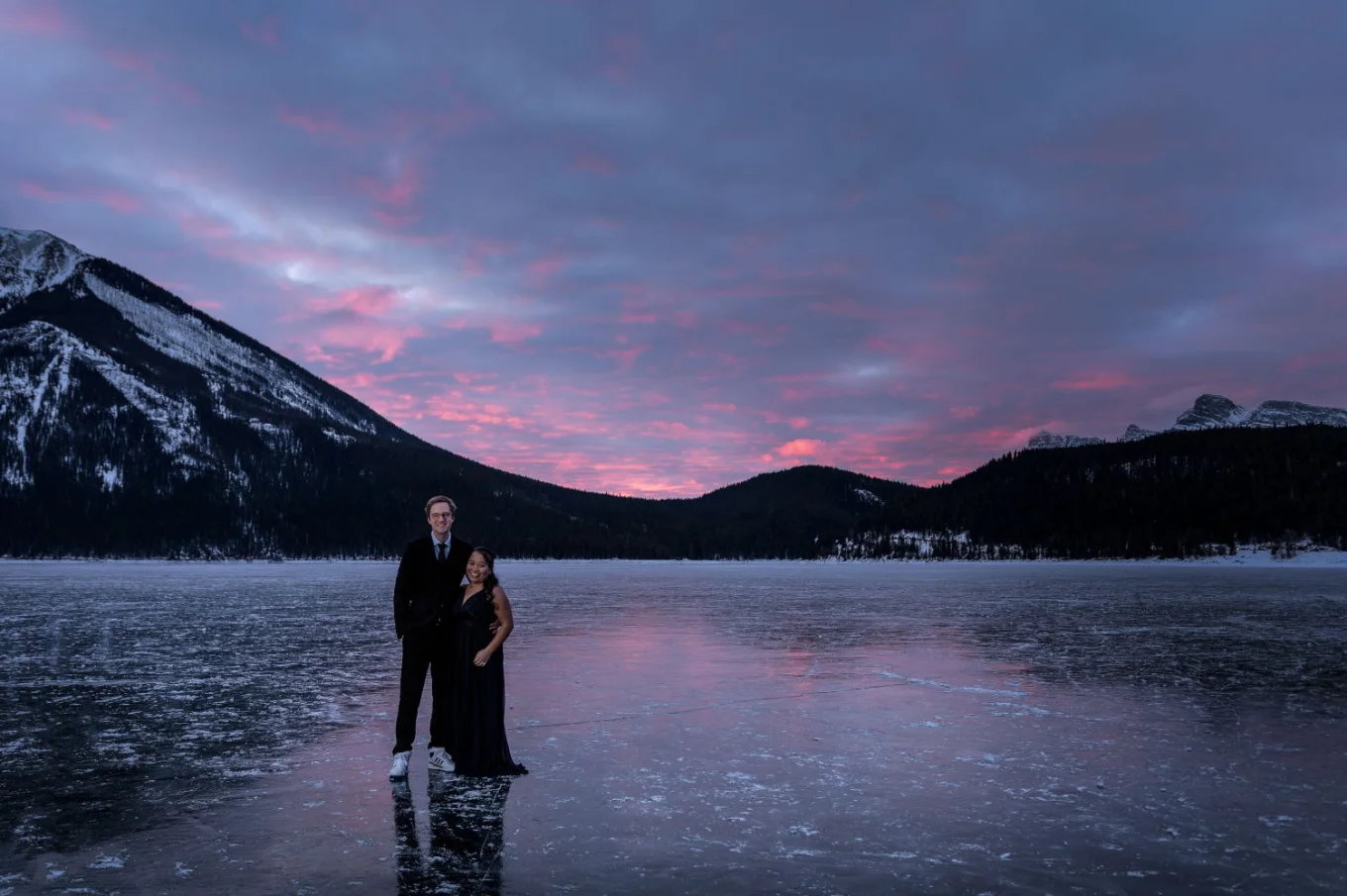 Stunning sunrise at Minnewanka Lake in Banff. Couple in love stand at the frozen lake an look at the photographer during photo session.