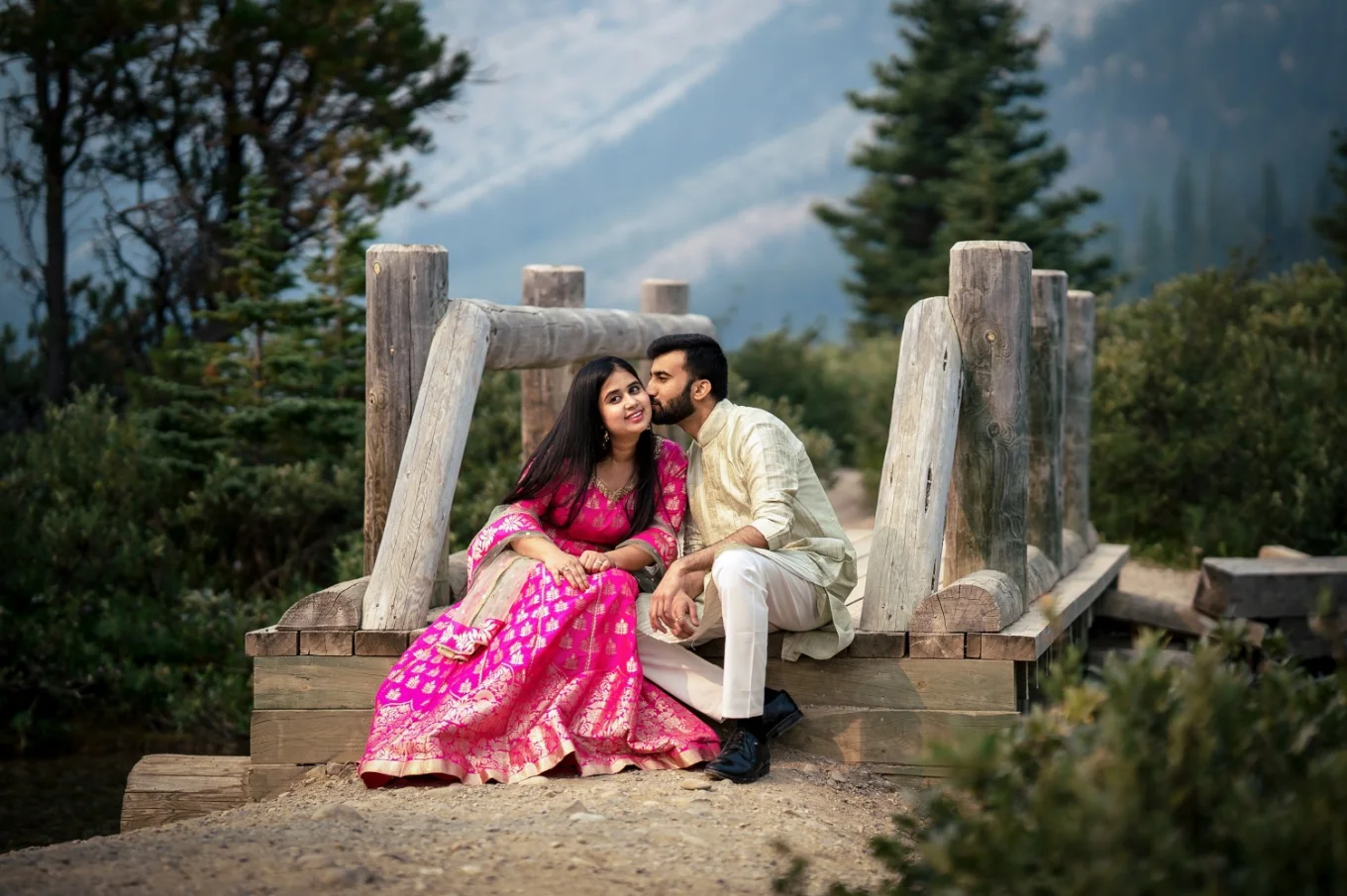 Indian couple wears traditional outfit. Man is kissing his fiancee on her cheek. They are sinning at the wooden bridge at Bow Lake.