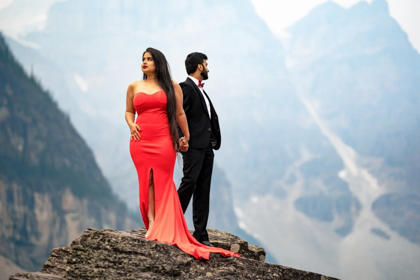 Lady in red dress look far away and her fiancee in suite look opposite direction. They are surrounded by the massive mountains in Banff.