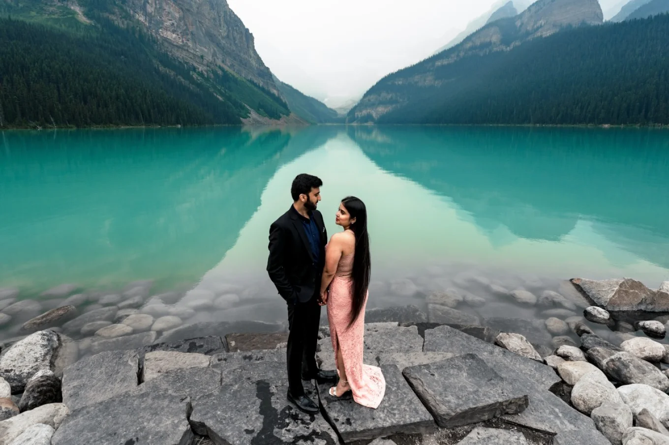 engagemant portrait an Lake Louise . The couple look at each other. Lady wears a pink dress and gentleman wears a black suite