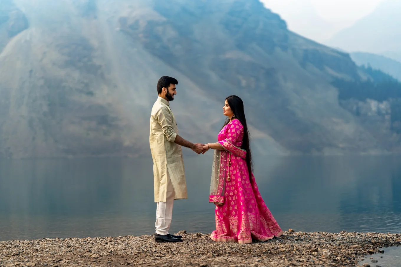 Indian couple wear traditional outfit. She wears a pink dress and he wears gold traditional outfit. They hold their hands and look at each other at Bow Lake in Banff