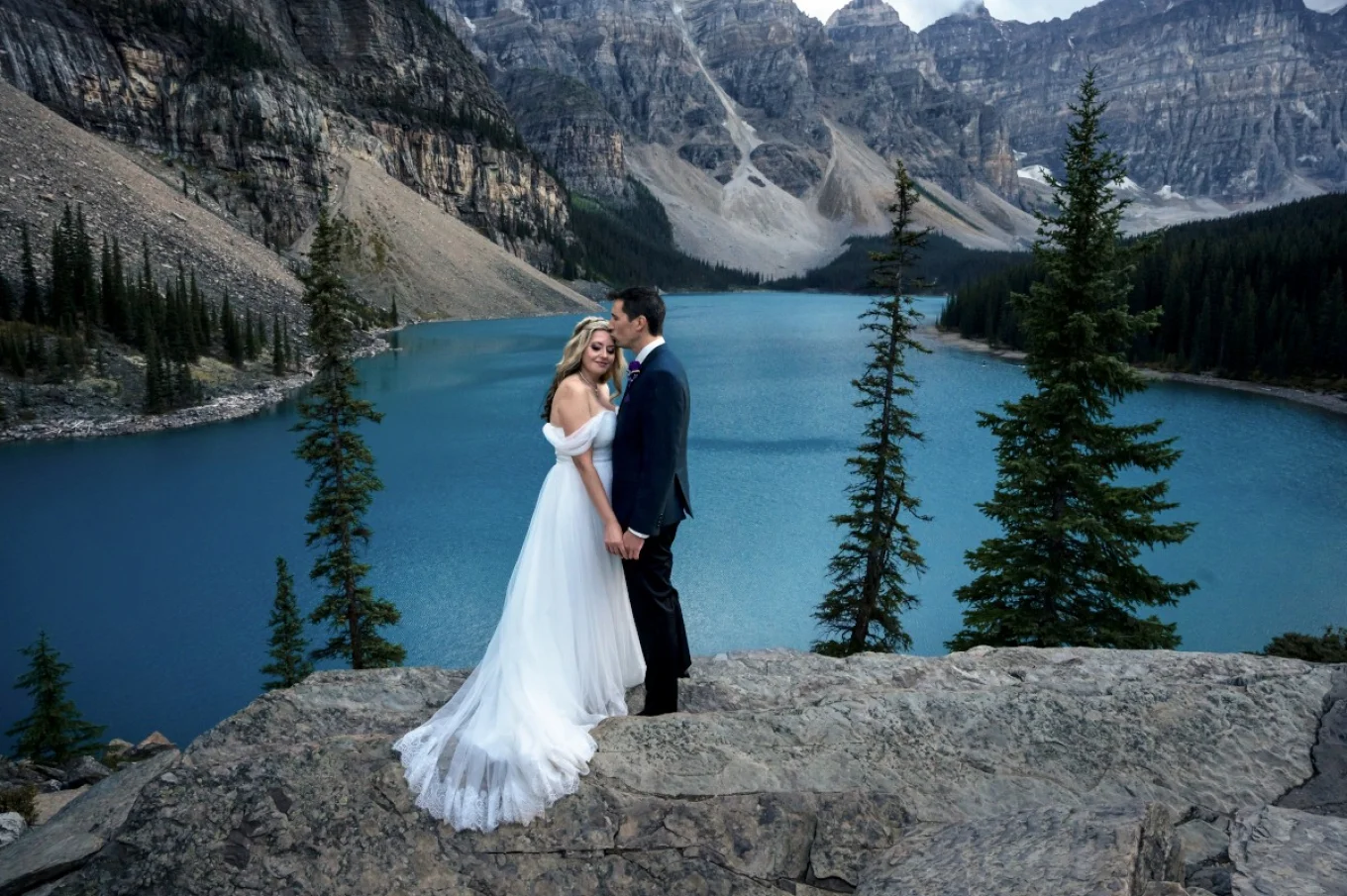 Groom kissing the bride at her forehead. They holds their hands at Moraine Lake in Banff.