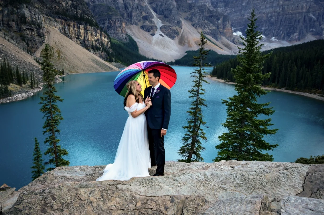 Bride and groom under colourful umbrella. They are surrounded by Canadian Rockies at Moraine Lake.