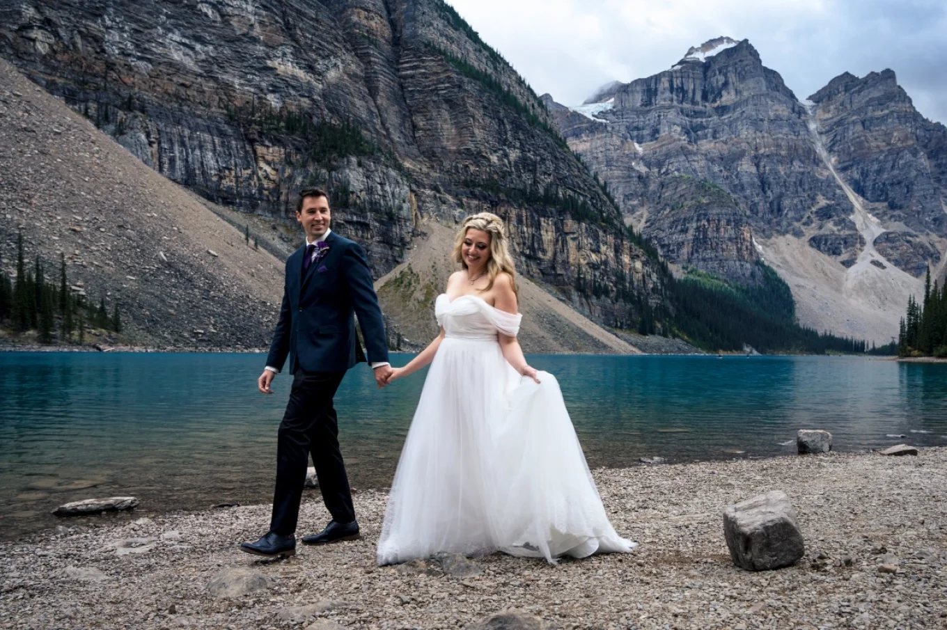 Bride and groom walk at Moraine Lake holding their hands. Groom is looking at his gorgeous wife.