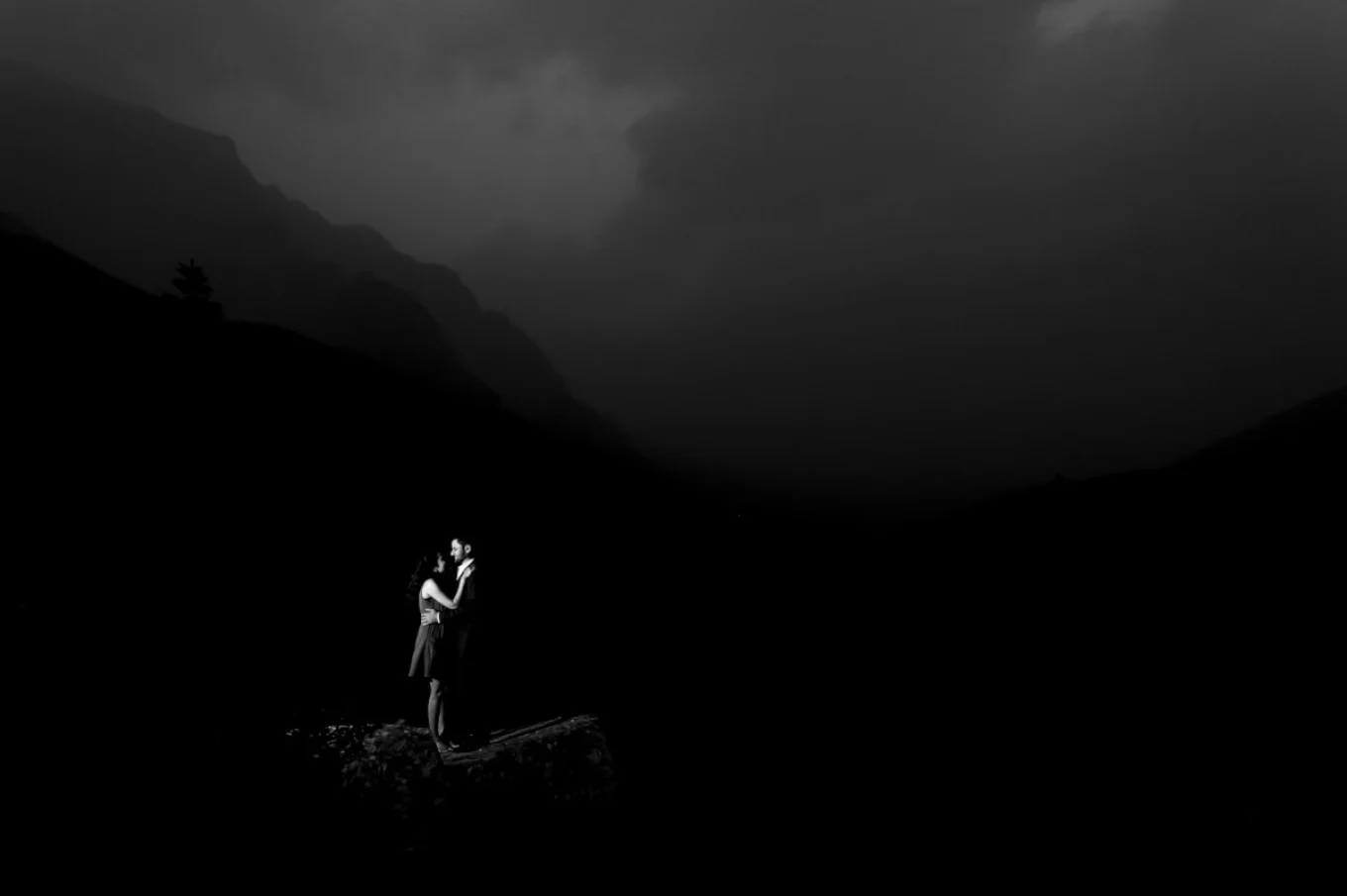 moody black and white engagement photo. Couple hug each other in the darkness