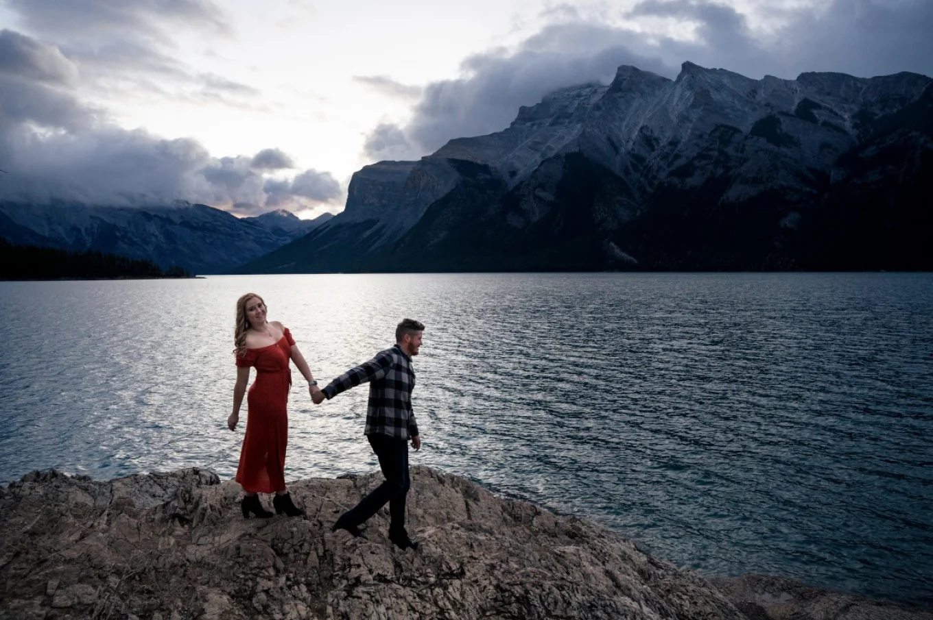 couple in love walking down the Minnewanka Lake in Banff. Their are at a huge rock and hold their hands.