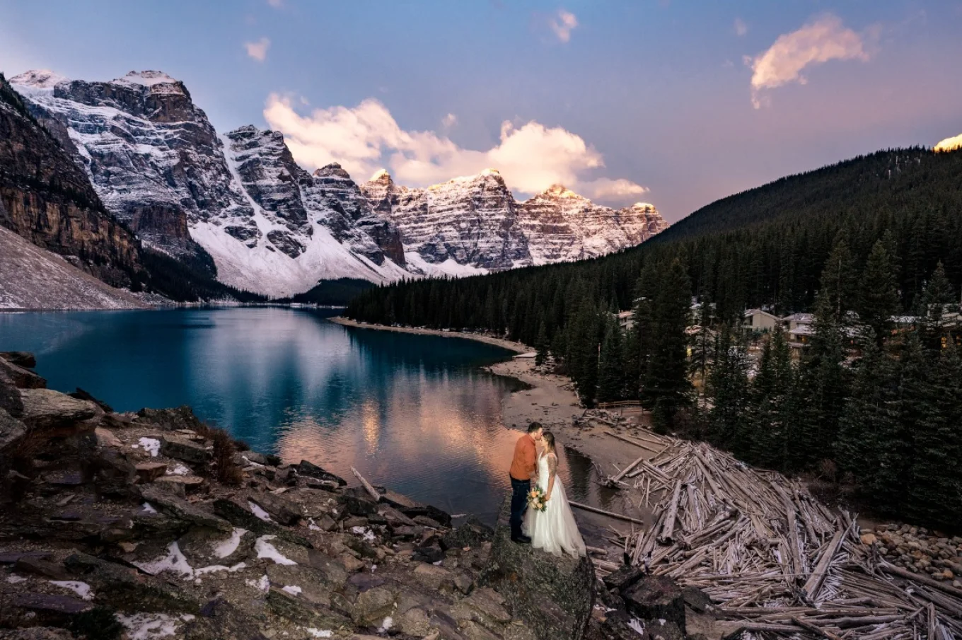 Newlyweds adore pink sunrise at Moraine Lake in Banff. They stand at the rock and look at the blue lake.