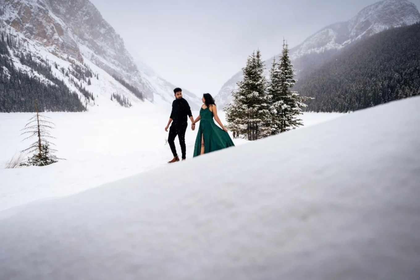 Couple in love walks down the hill at Lake Louise in wintertime. She wears green, long dress, he is dressed n black.