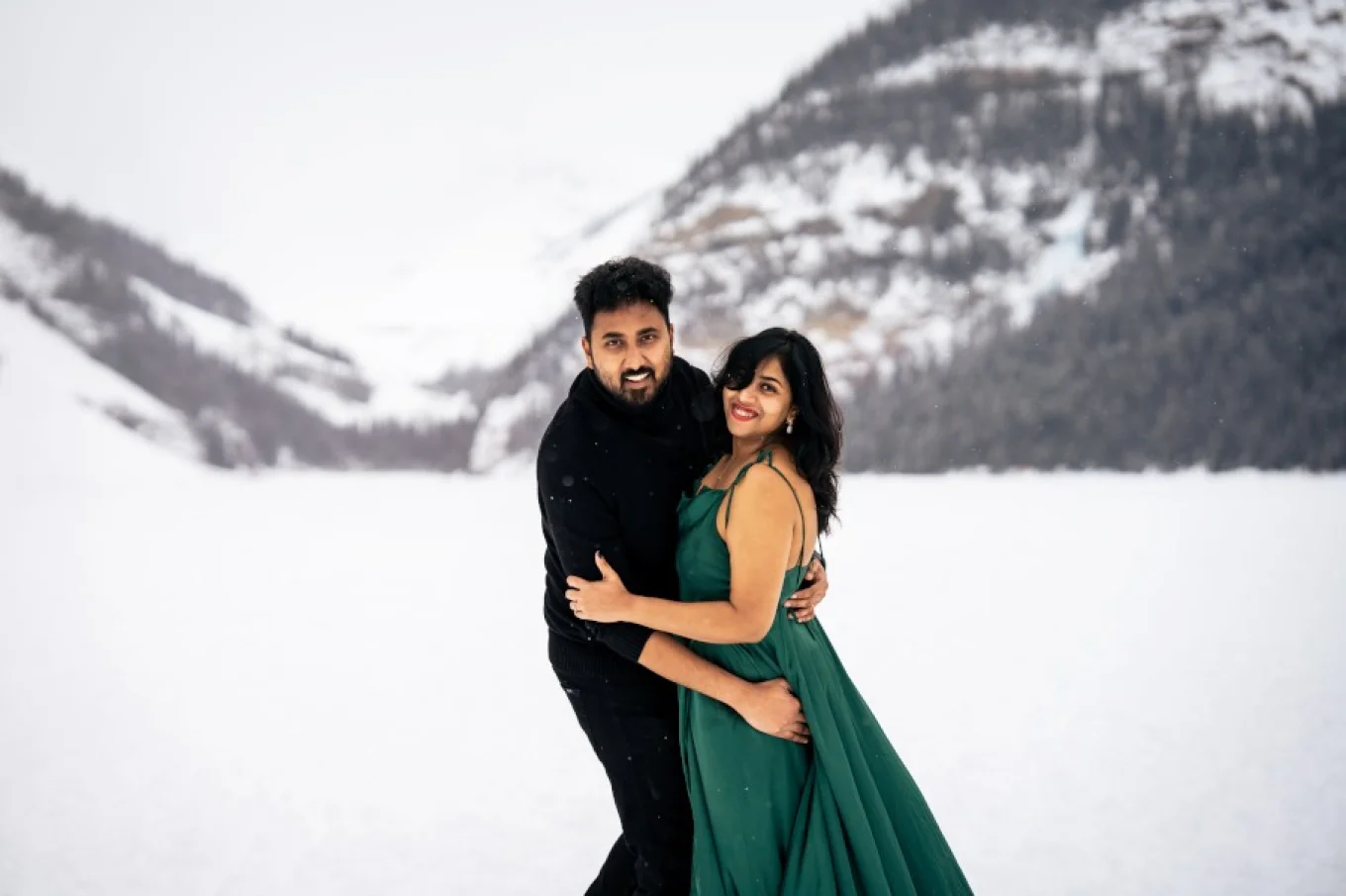 Indian couple smiling and looking at the calera of Calgary Wedding Photographer. Winter photo session at Lake Louise in Banff.