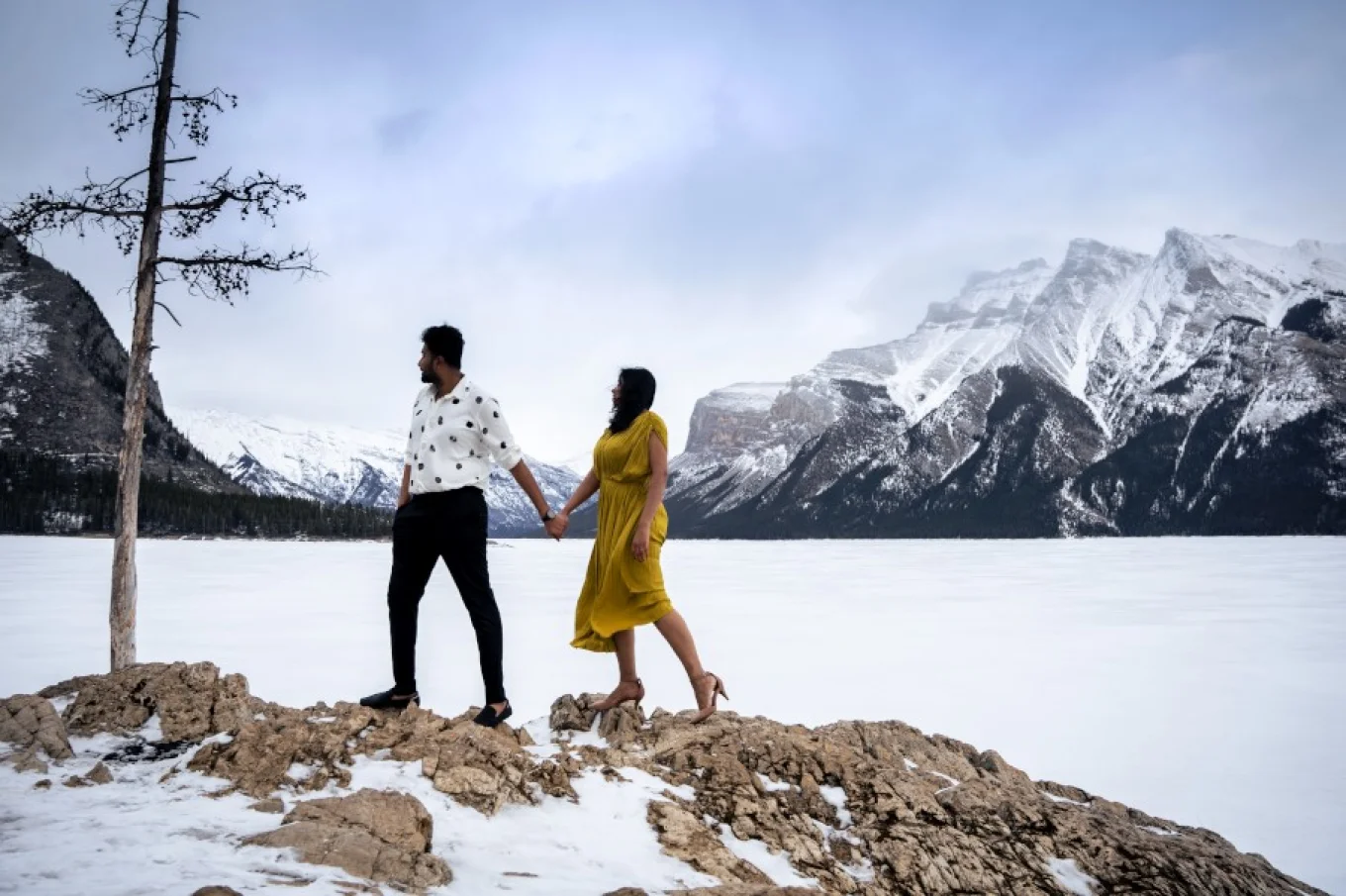 Man and woman stand at the edge of the frozen Minnewanka Lake. They are surrounded by Canadian Rocky Mountains.