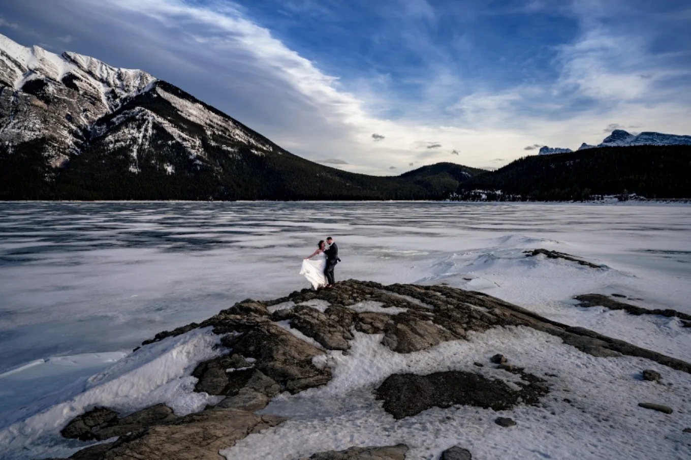 After wedding session at Minnewanka Lake with the best photographer Eliza Slobodzian. Couple stands at the frozen lake. They are surrounded by the Canadian Rockies.
