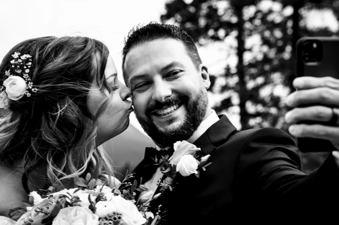 Bride kisses cheek of her husband. The broom takes selfie of them.