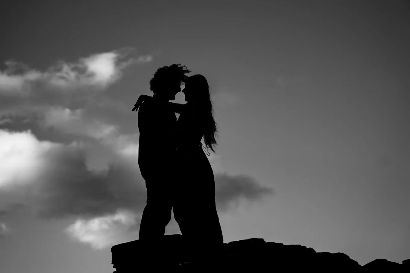 silhouette photograph of engaged couple in Black and white. They hug each other on the rock. In the background is sky.