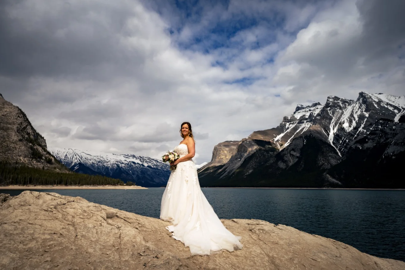 Bride's portrait. She stands on the big rock by the Minnewanka Lake. She holds bouquet and smiles.