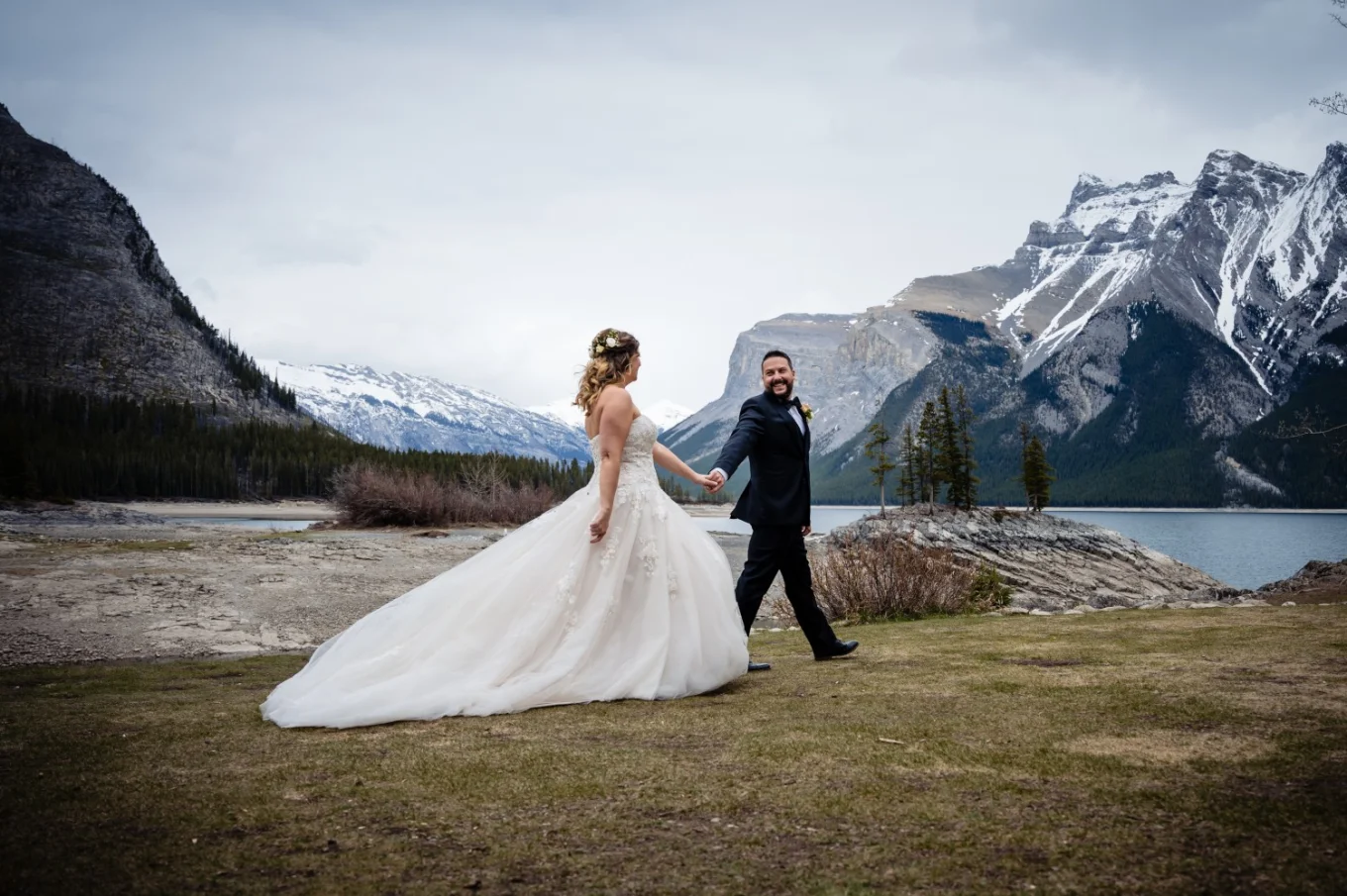 Bride and groom walk down the Minnewanka Lake . They hold their hands and look at each other.