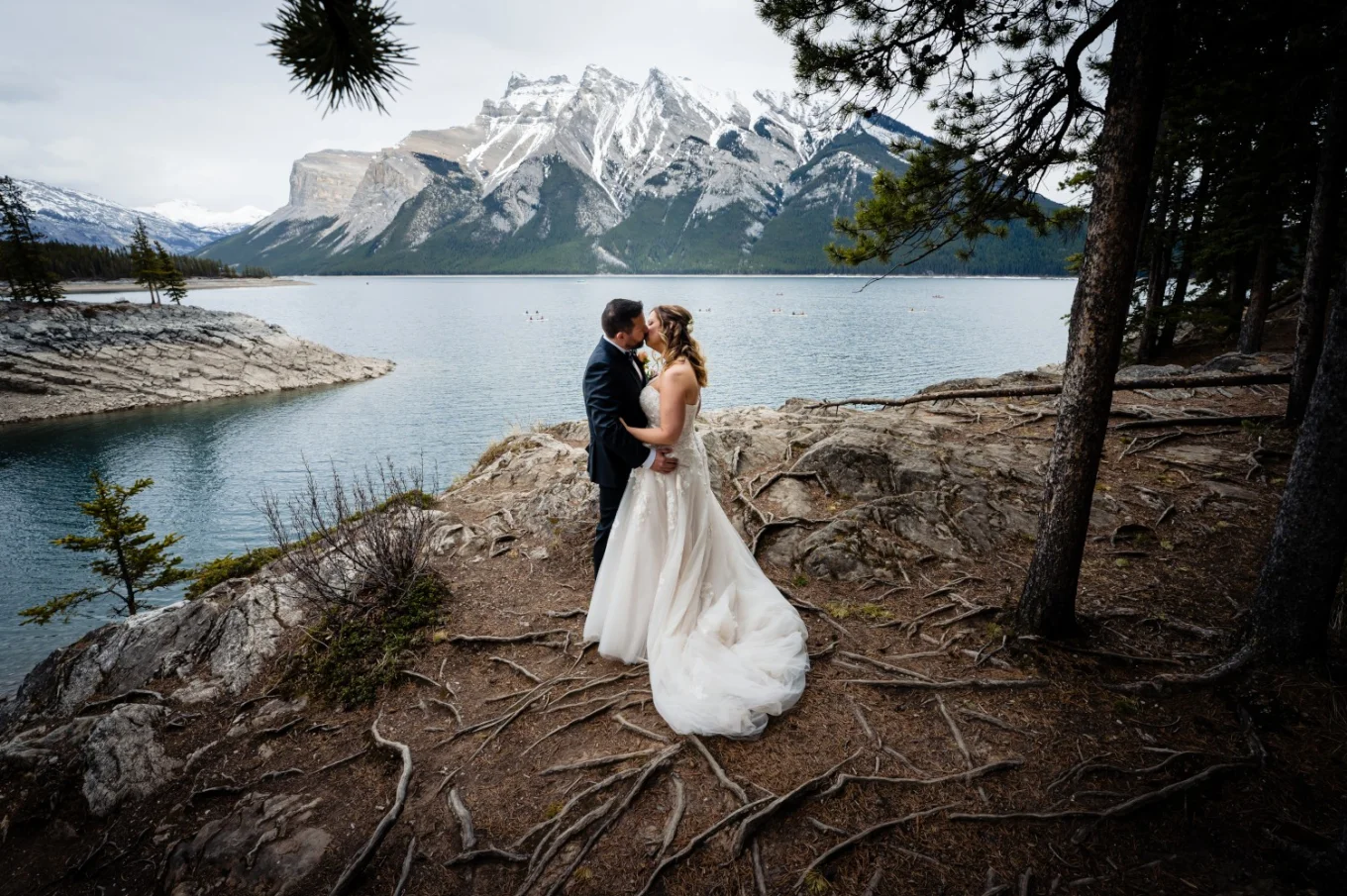 Newlyweds kiss during their wedding photo session at Minnewanka Lake. On the background big mountain covered with snow.