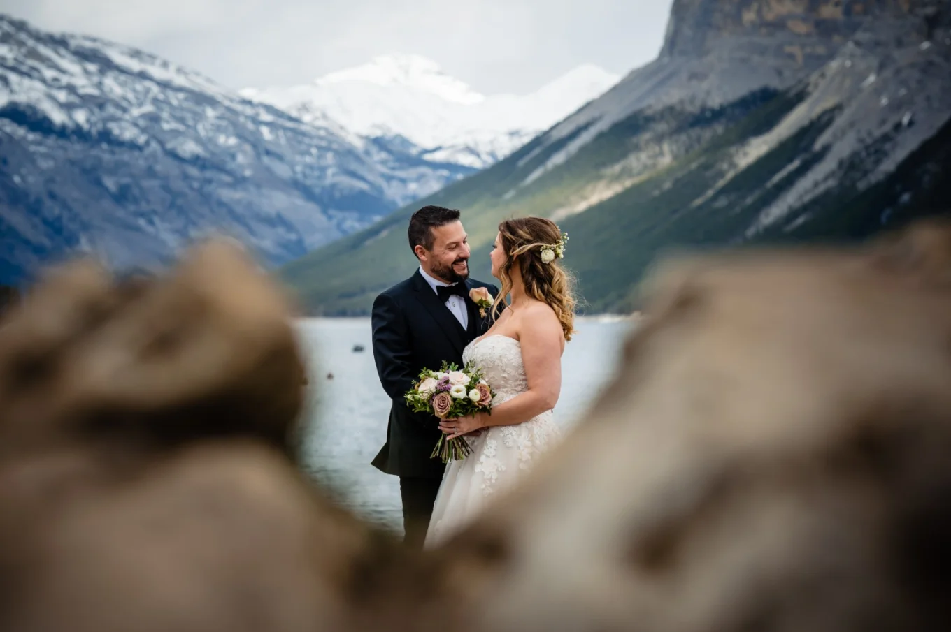 bride and groom look at each other and hug. Stunning mountains and blue Minnewanka Lake on the background. They ate kept in the frame of the rocks.