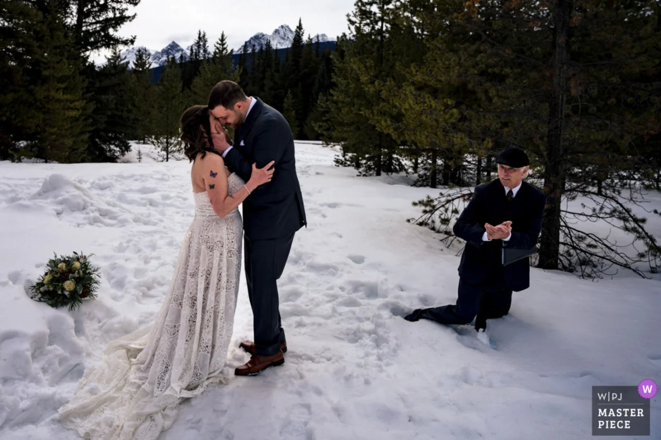 First kiss of bride and groom during Banff elopement. Officiant clapping hands fall down into deep snow.