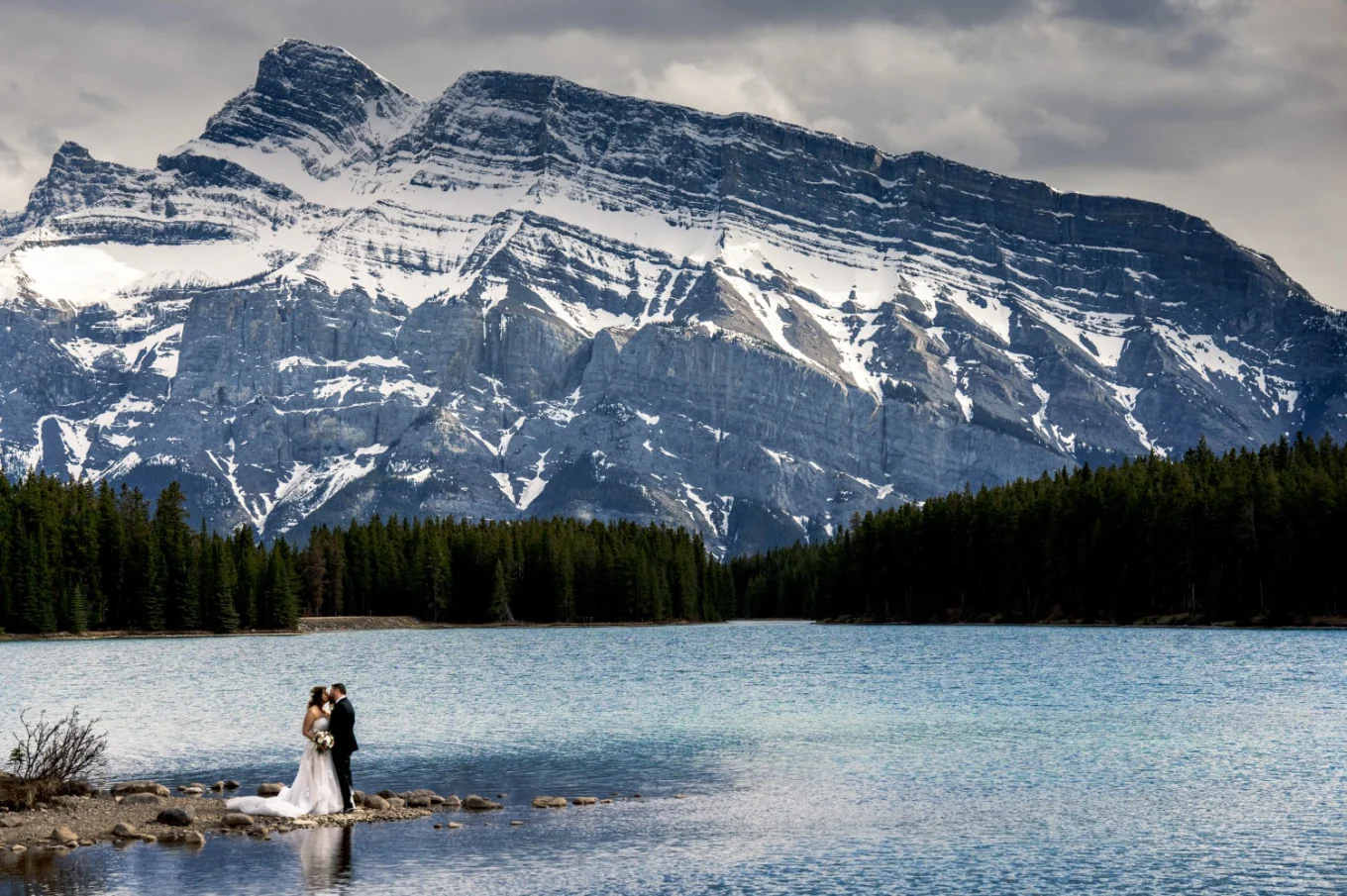 Newlyweds hug during wedding photo session captured by photographer Eliza Slobodzian. Couple is surrounded by a giant mountain and Two Jack Lakes in Banff.