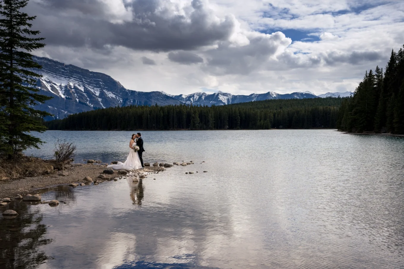 Newlyweds standing at the edge of Two Jack Lakes in Banff and hug. Amazing Canadian Rockies on the background.