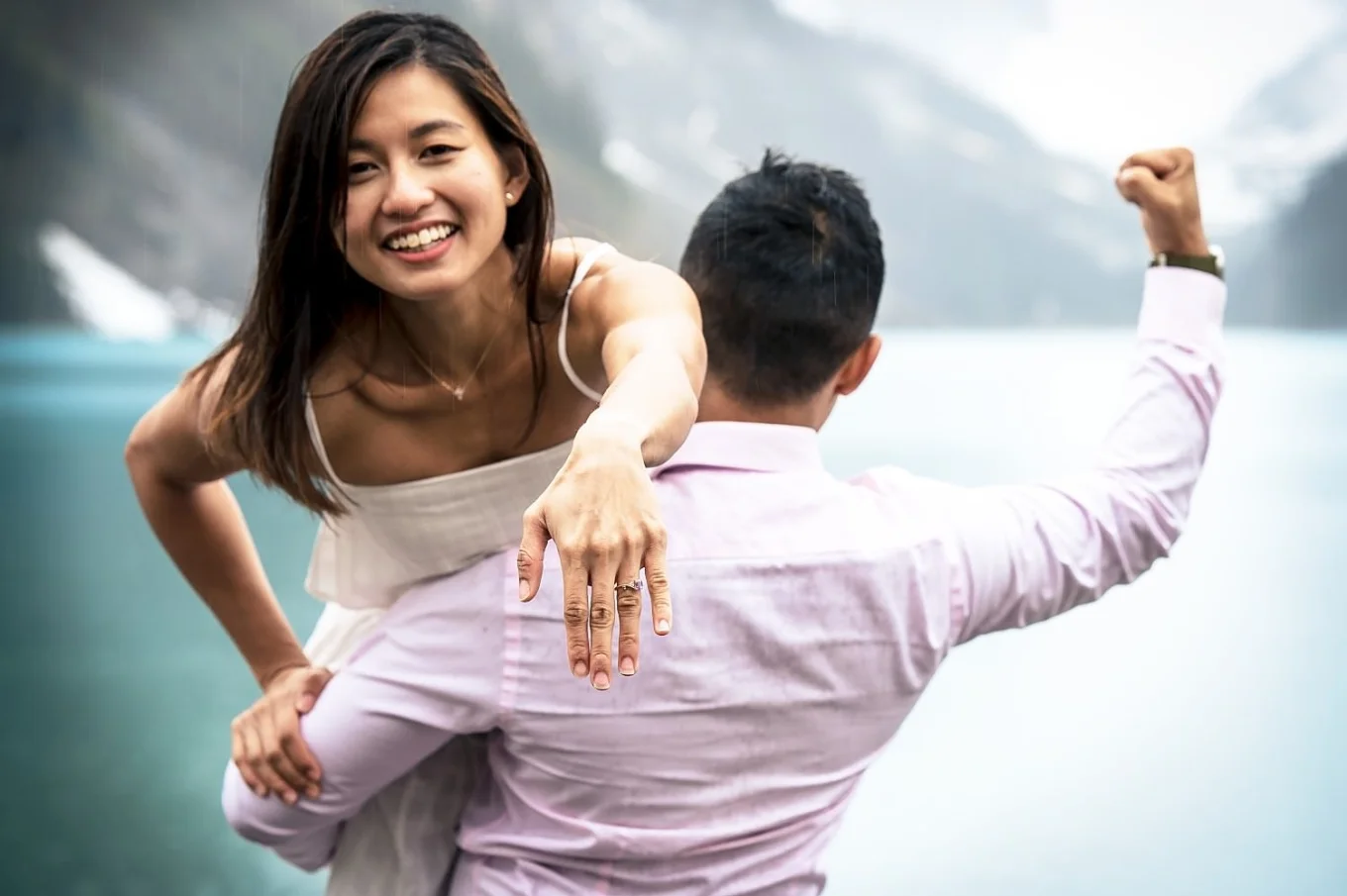 Man carry his fiancee on his shoulder while she laugh and shows her hand with an engagement ring.