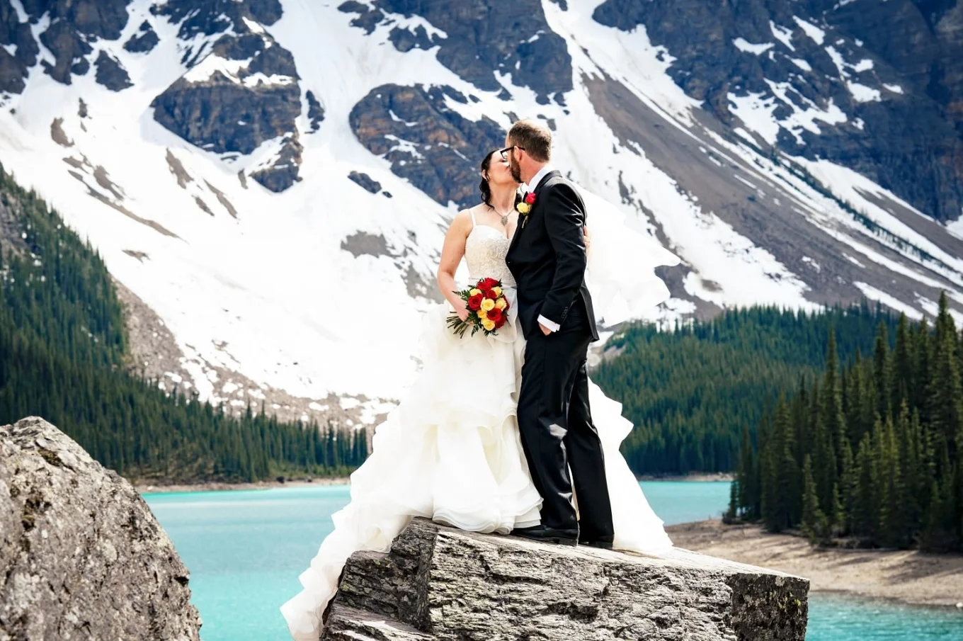 Newlyweds share a kiss at Moraine Lake in Banff. They are surrounded by snowy mountains.