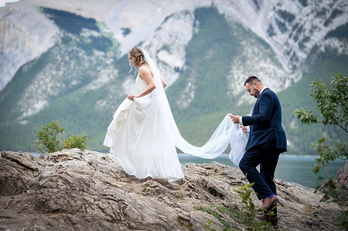 Groom helps the bride with her veil while climbing the giant stone at Minnewanka Lake.