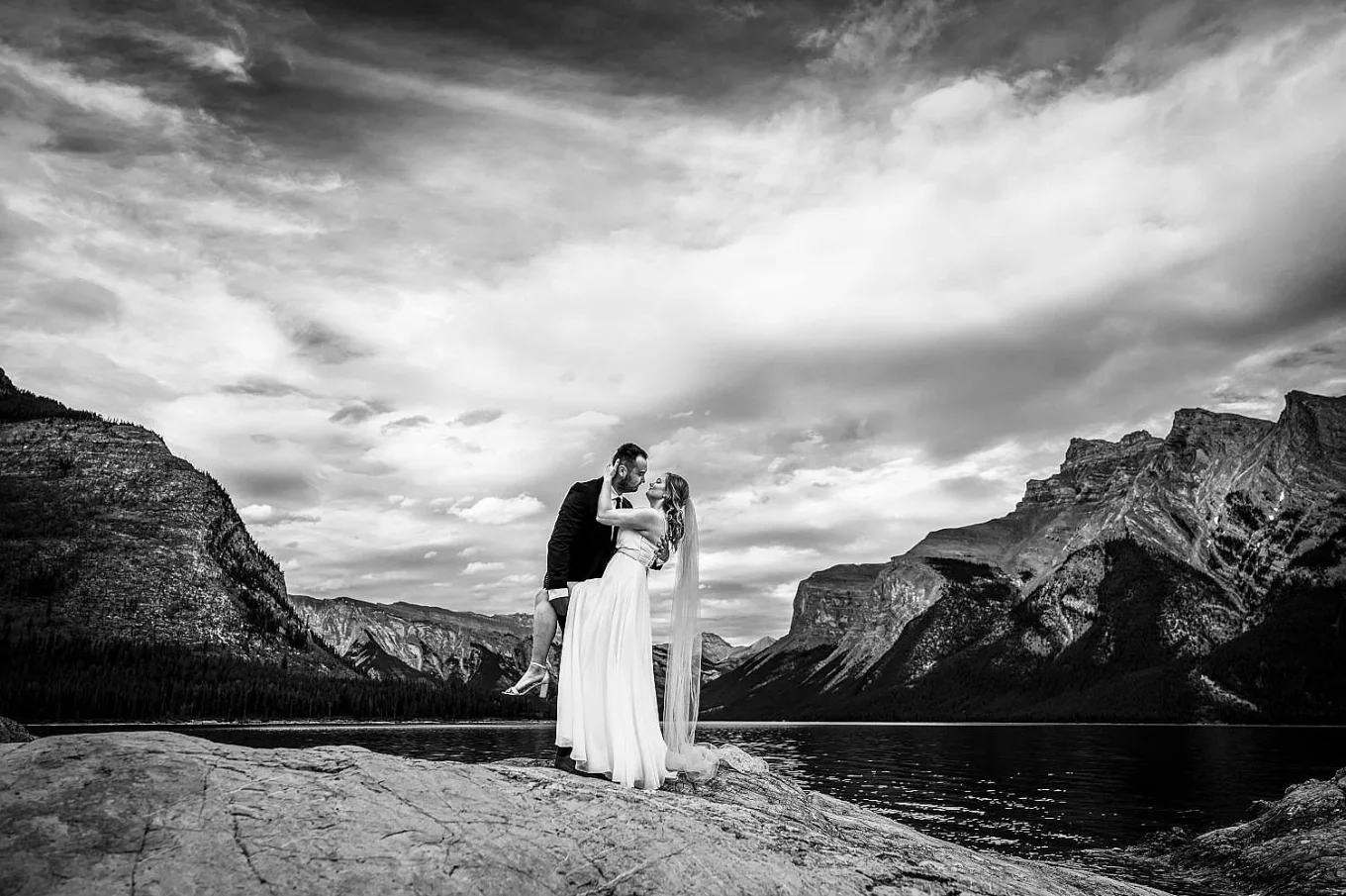black and white photograph in the Canadian Rockies. Bride and groom dance by Minnewanka Lake in Banff.