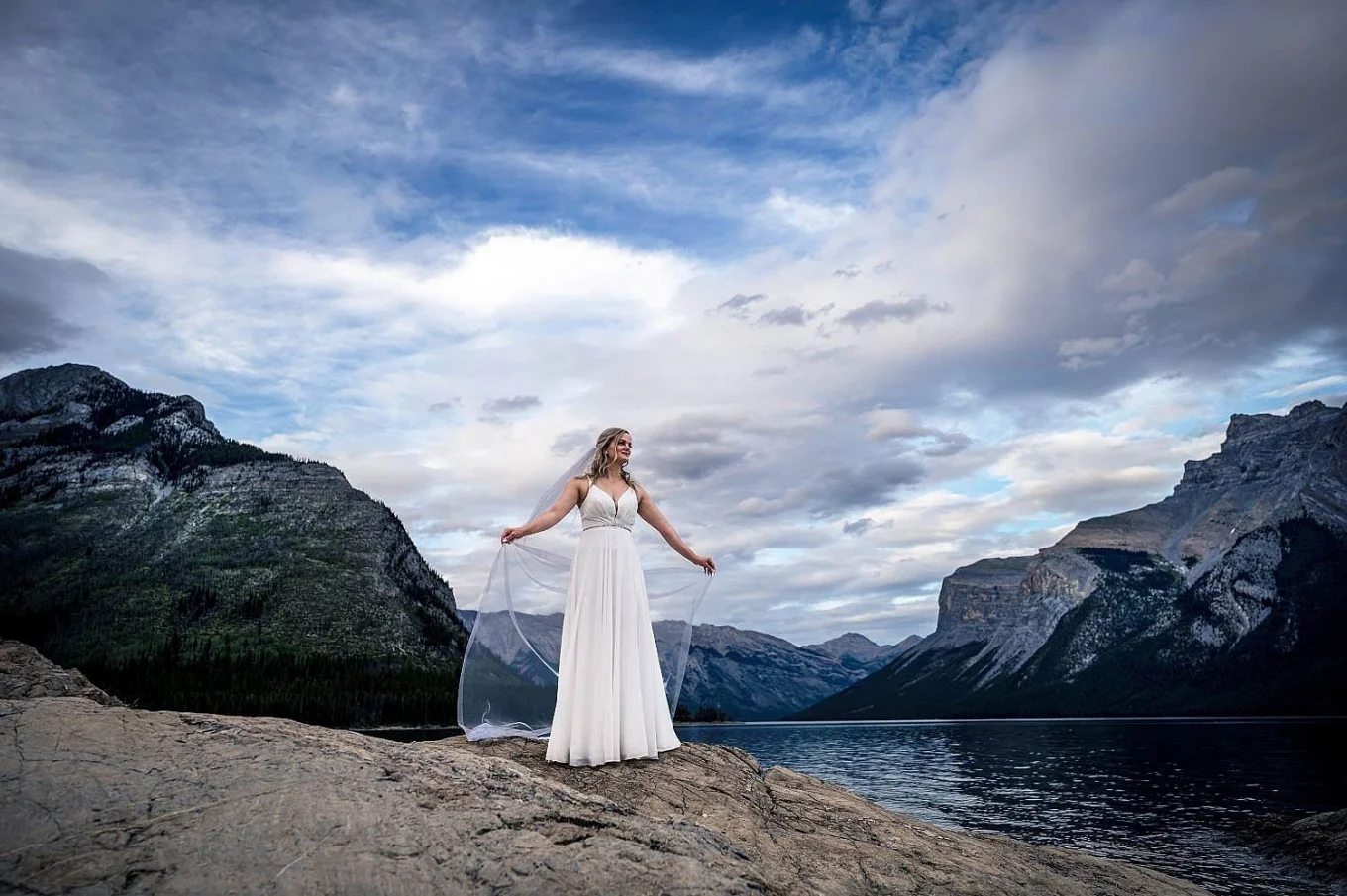 Amazing bride's portrait at Minnewanka Lake captured by Banff Wedding Photographers. Bride holds her veil and look at the lake.