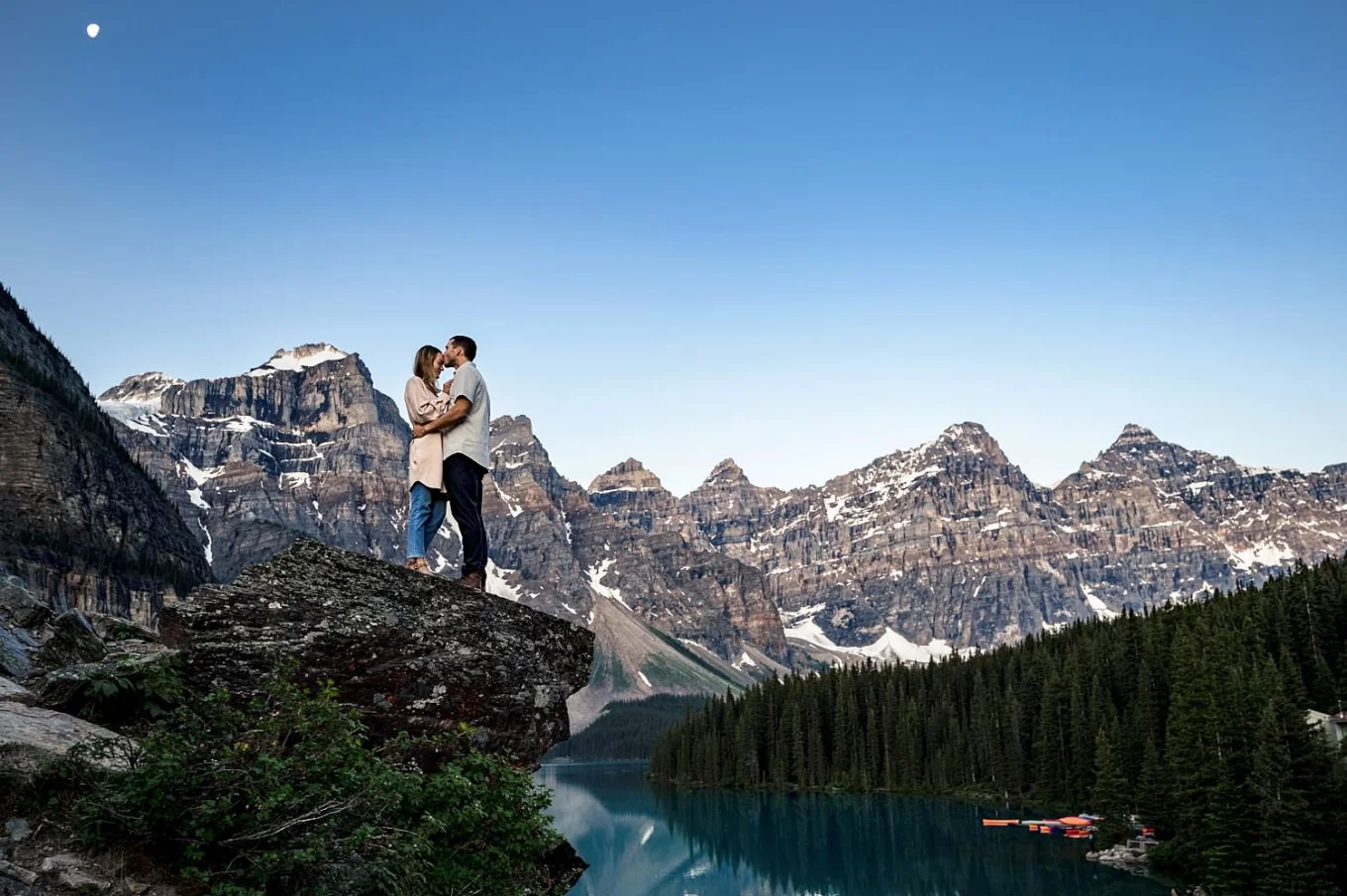 Newly engaged couple in love hugging during their engagement session at Moraine Lake. They stand at the edge ot the rock against tremendous mountains and lake/