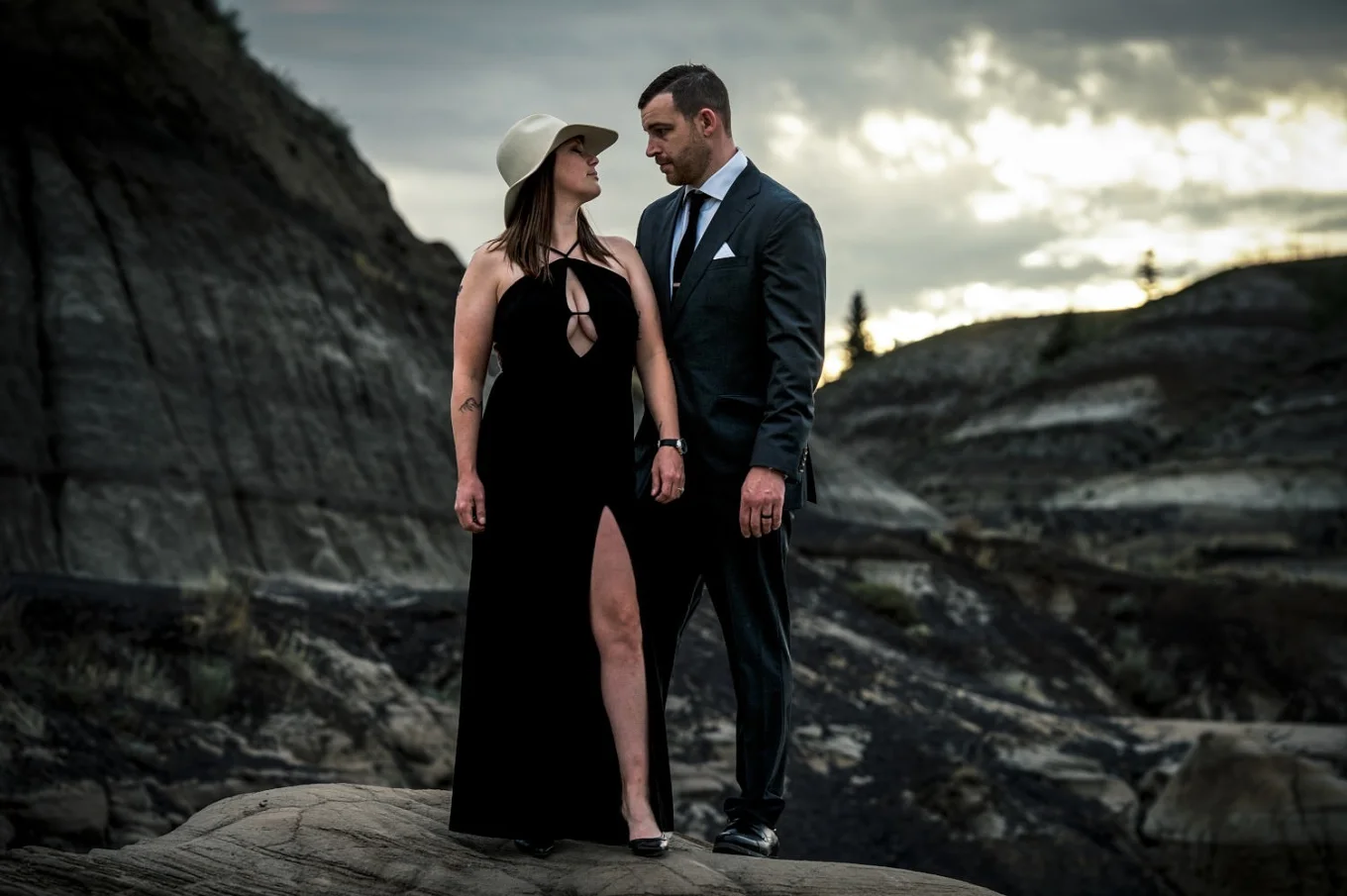 Married couple during after-wedding session. lady wears log black dress and a hat and Gentleman wears suit. They loos at each other with love.