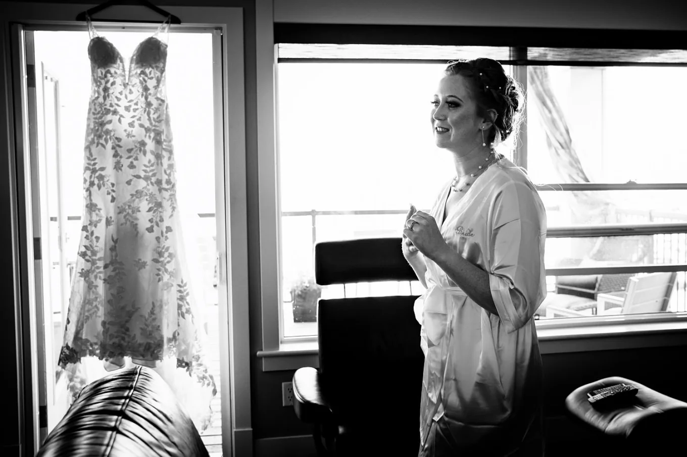 Photograph of the bride wearing a robe and a wedding dress hanging on the window.