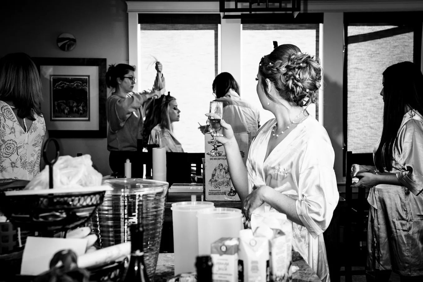 Black and white photograph during Calgary wedding. Bridesmaid having a hair done and the bride look at her with a glass of champagne in her hand.