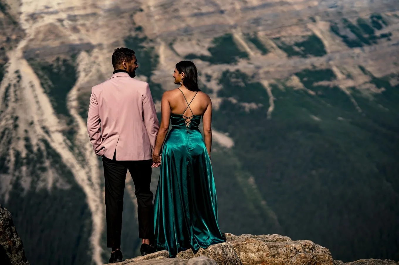 A man in a suite and a lady in a long, green dress from behind. They look at each other.