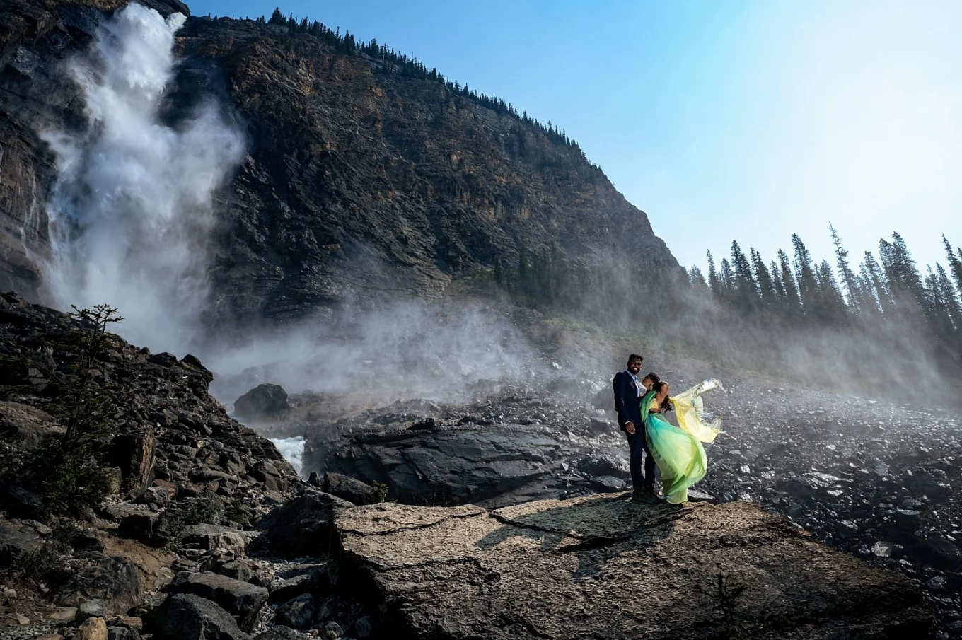 Engagement photograph of couple in a hug at Takakkaw Falls. Wind is blowing the girl yellow dress.