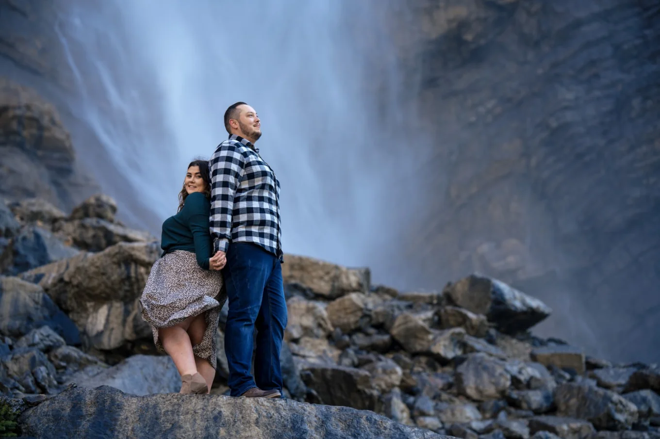 Engaged couple standing back to back and holding hands against breathtaking Takakkaw Falls in Banff.