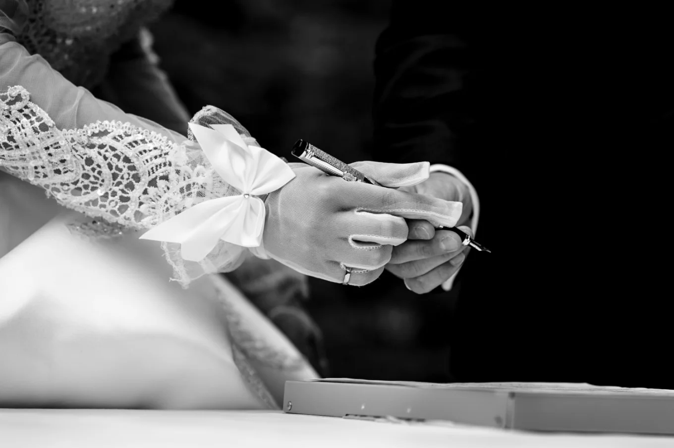 Black and white photograph of newlyweds hands. Bride passing a pen to the groom.