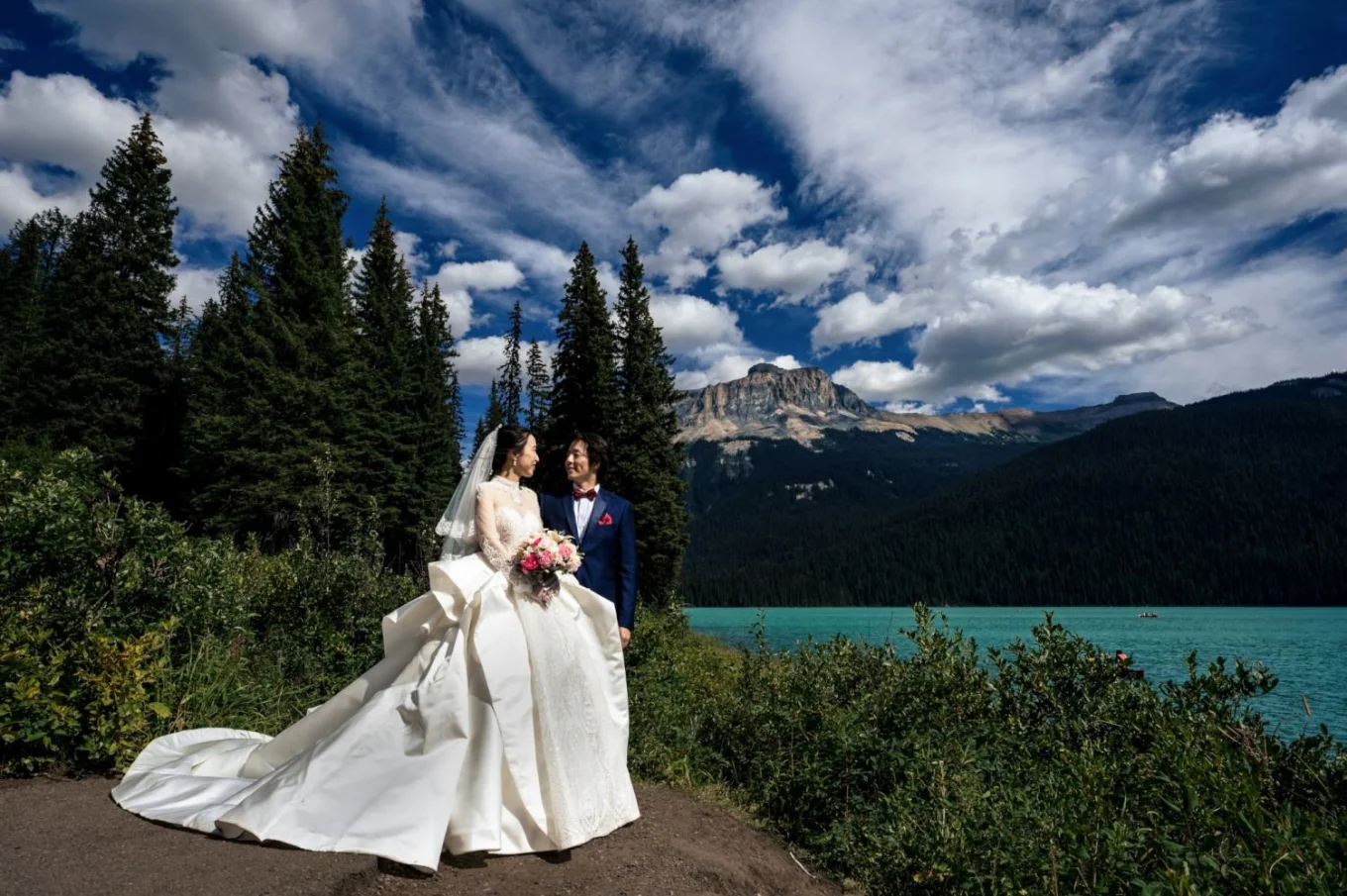 Newlyweds portrait at Emerald Lake . Behind the couple tall trees , lake and the mountains. Above their heads interesting sky with moving clouds,