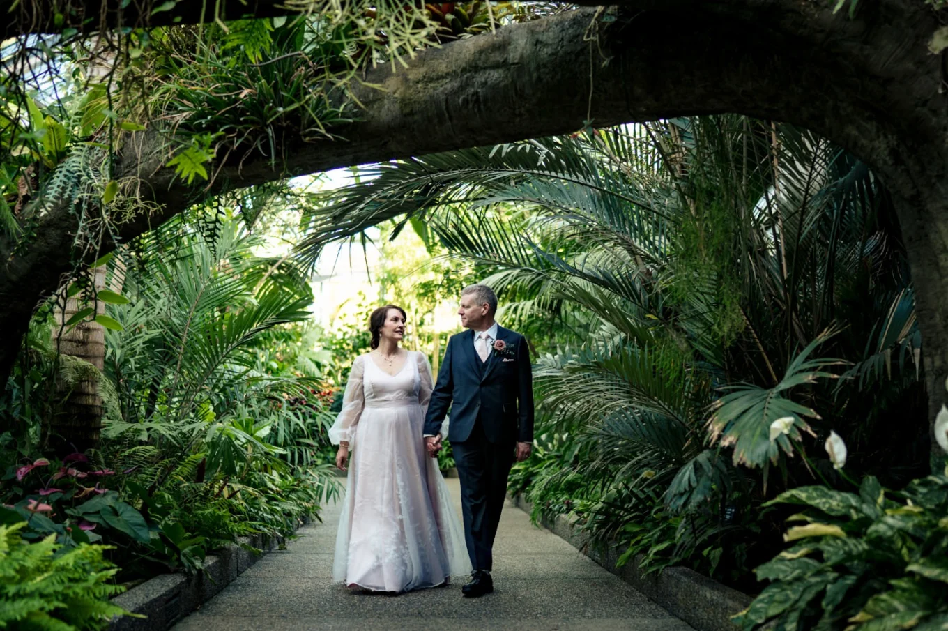 Newlyweds walk in the Calgary Zoo garden, holding hands and looking at each other.