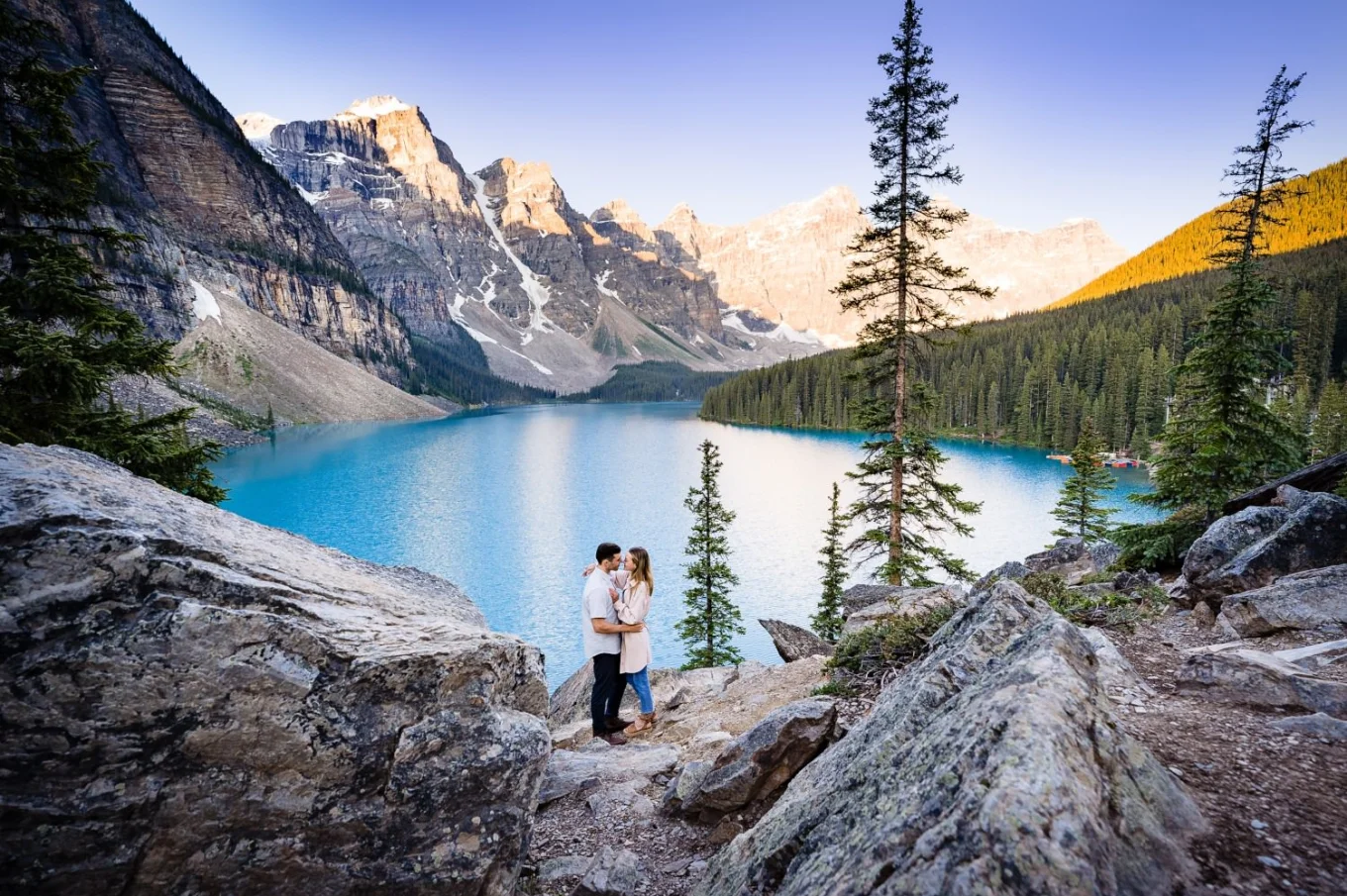 Engagement young couple posing to Banff Wedding Photographer at amazing location of Moraine Lake. They are in a hug, facing each other. Behind them beautiful lake and huge mountains